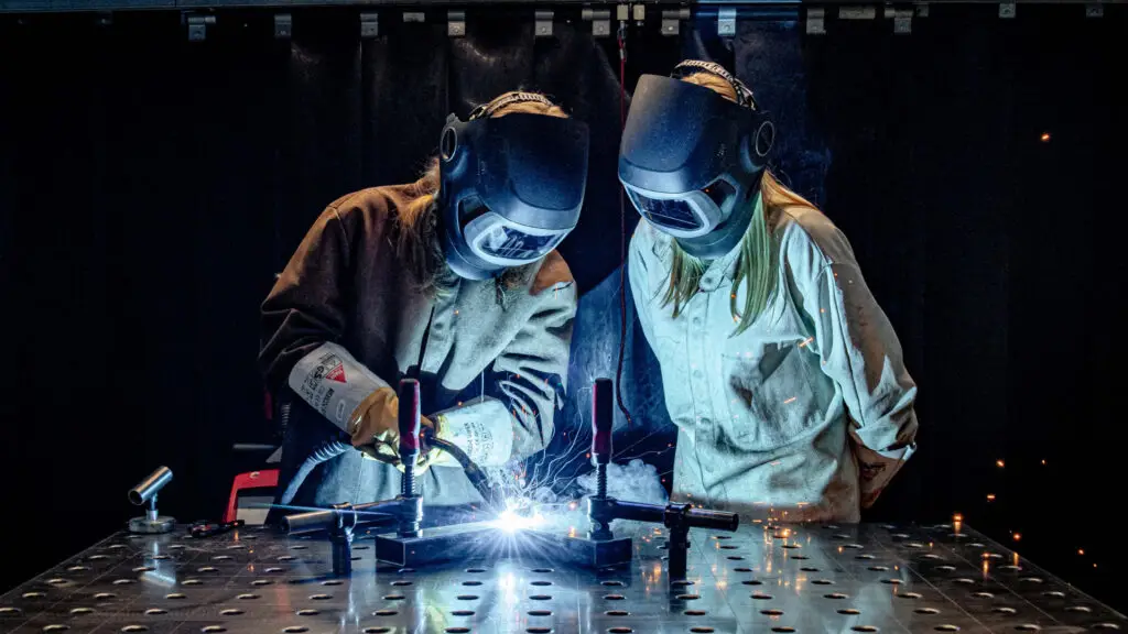 Aufnahme von zwei Personen mit langen Haaren und Schweißerhelm beim Schweißen von Metall