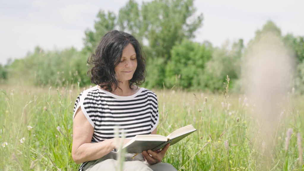 Eine schwarzhaarige Frau mit einem schwarz weiß gestreiftem Oberteil, die in einer Wiese sitzt und ein Buch ließt